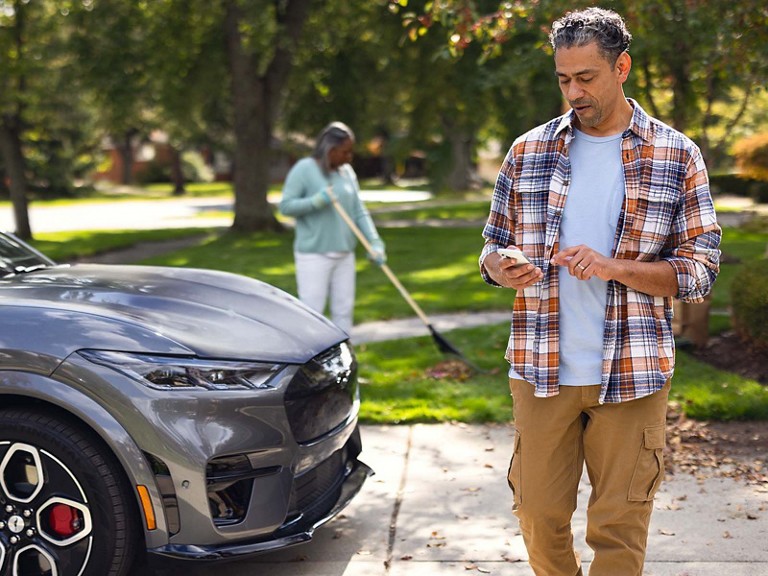 An owner strolls around their Mustang Mach-E® in the driveway scrolling on their phone