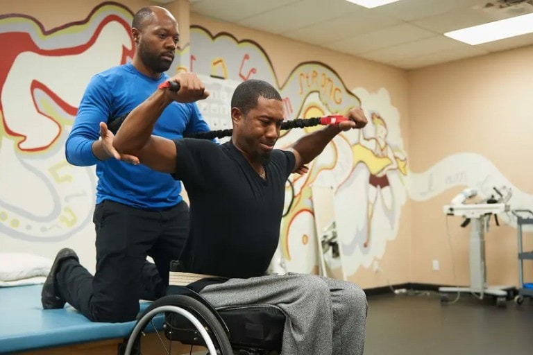 A physical trainer is kneeling behind a man in a wheelchair at a rehabilitation center, taking him through some resistance training with a resistance band