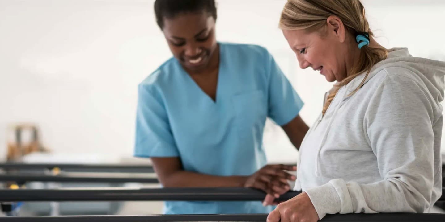 A nurse in scrubs helps a woman to walk between balance bars in a rehabilitation center