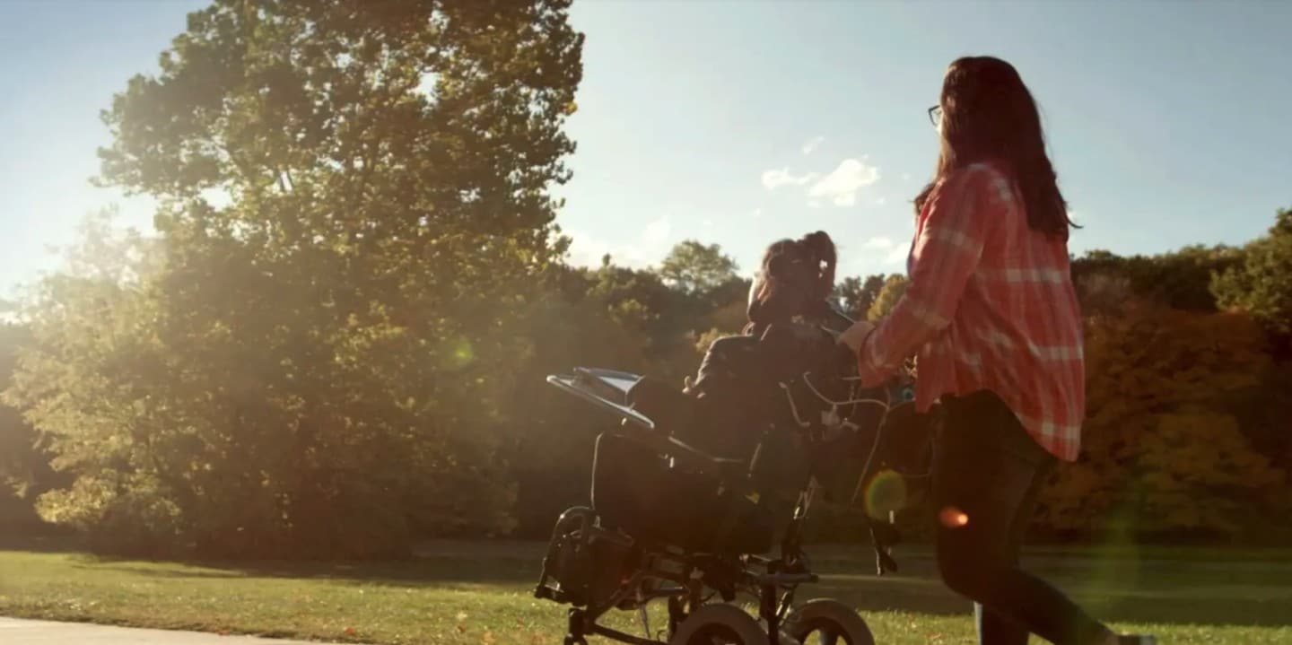A woman is walking along the sidewalk pushing her daughter Kylie in her wheelchair.
