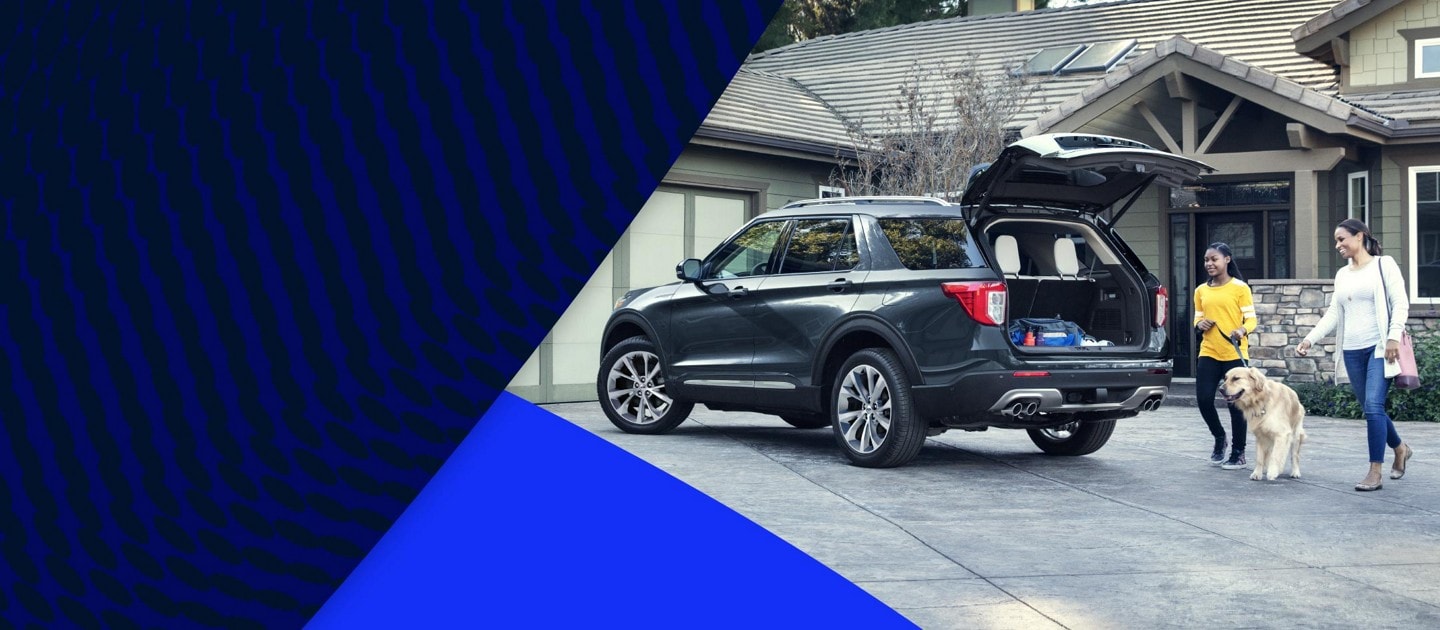 Two people and a dog approach the rear open hatch of a Ford Explorer® SUV parked in a driveway