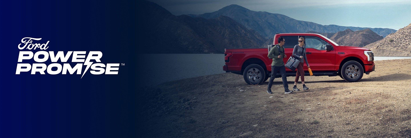 A couple walking next to a red F-150 Lightning near a sandy lake with mountains in the background