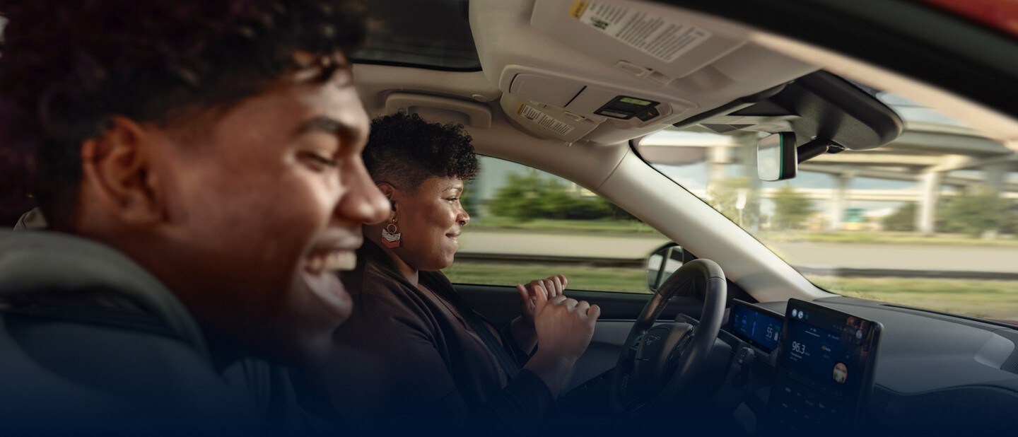 A mother drives a 2023 Ford Mustang Mach-E with her hands off the steering wheel and her teenage son in the passenger seat