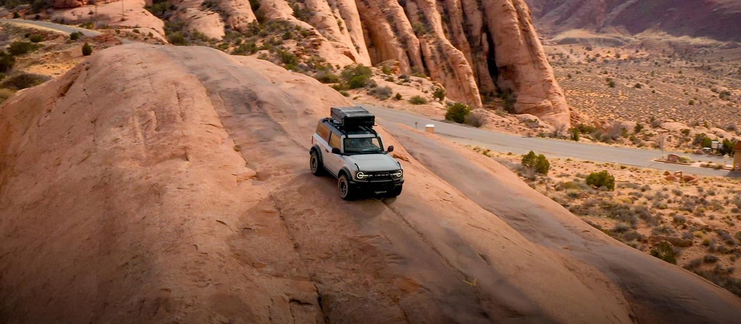A gray two-door 2023 Ford Bronco® drives down a rock formation with a cargo carrier attached to its roof