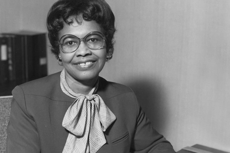 We see a black and white portrait of auto innovator Dr. Gladys West sitting at her desk.