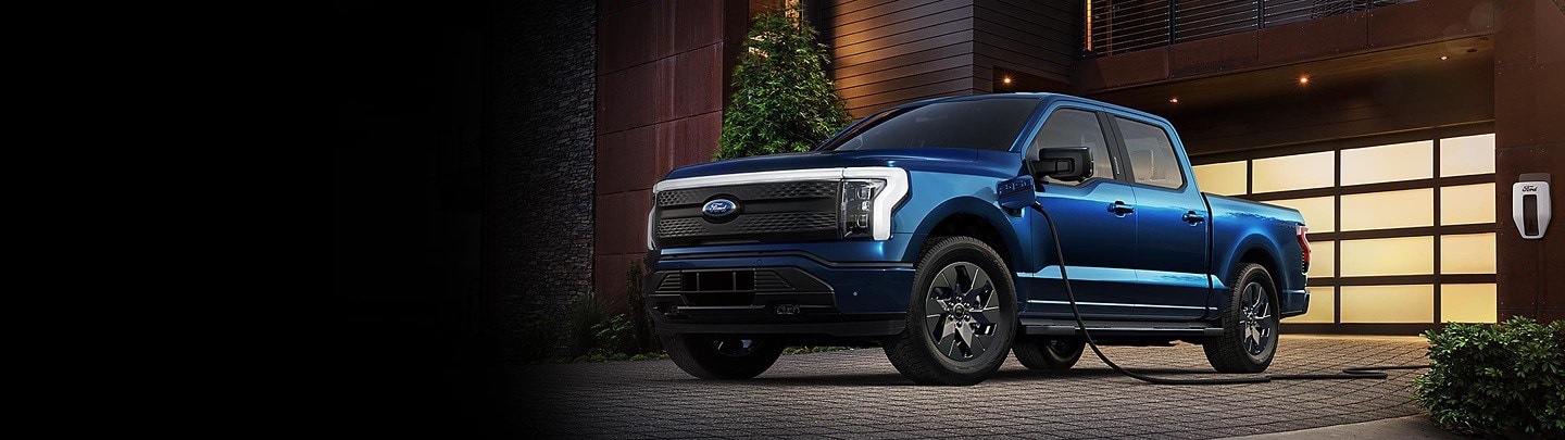 A blue Ford F-150® Lightning® parked in a driveway at night. It is plugged into a Ford Charge Station Pro