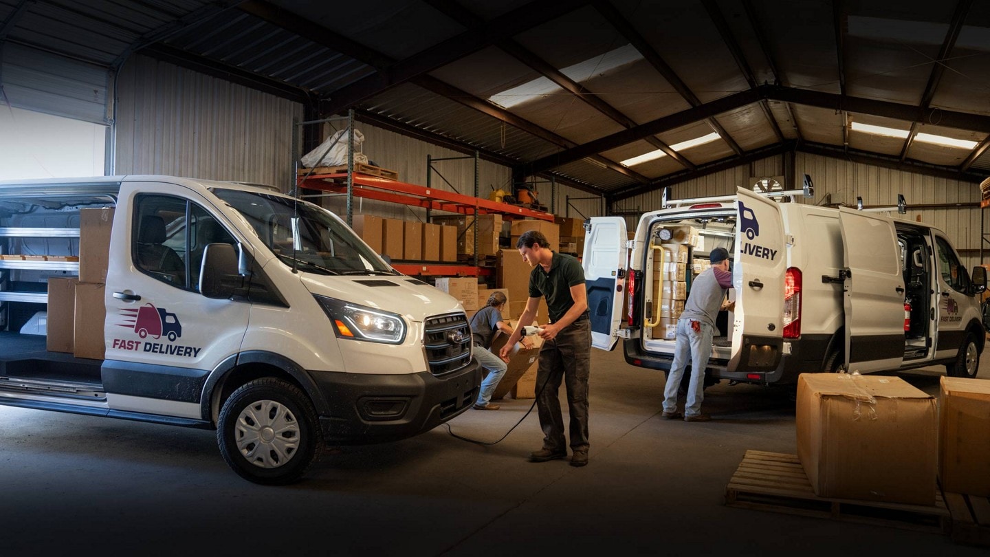 A pair of white 2024 Ford E-Transit™ vans parked in warehouse. One worker plugs in a charge cable while another loads cargo
