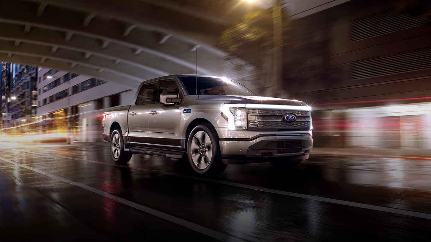 A silver 2024 Ford F-150 Lightning® pickup truck driving down a rainy city street at night