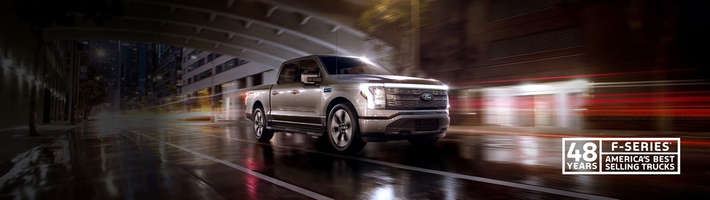 A silver 2024 Ford F-150 Lightning® pickup truck driving down a rainy city street at night