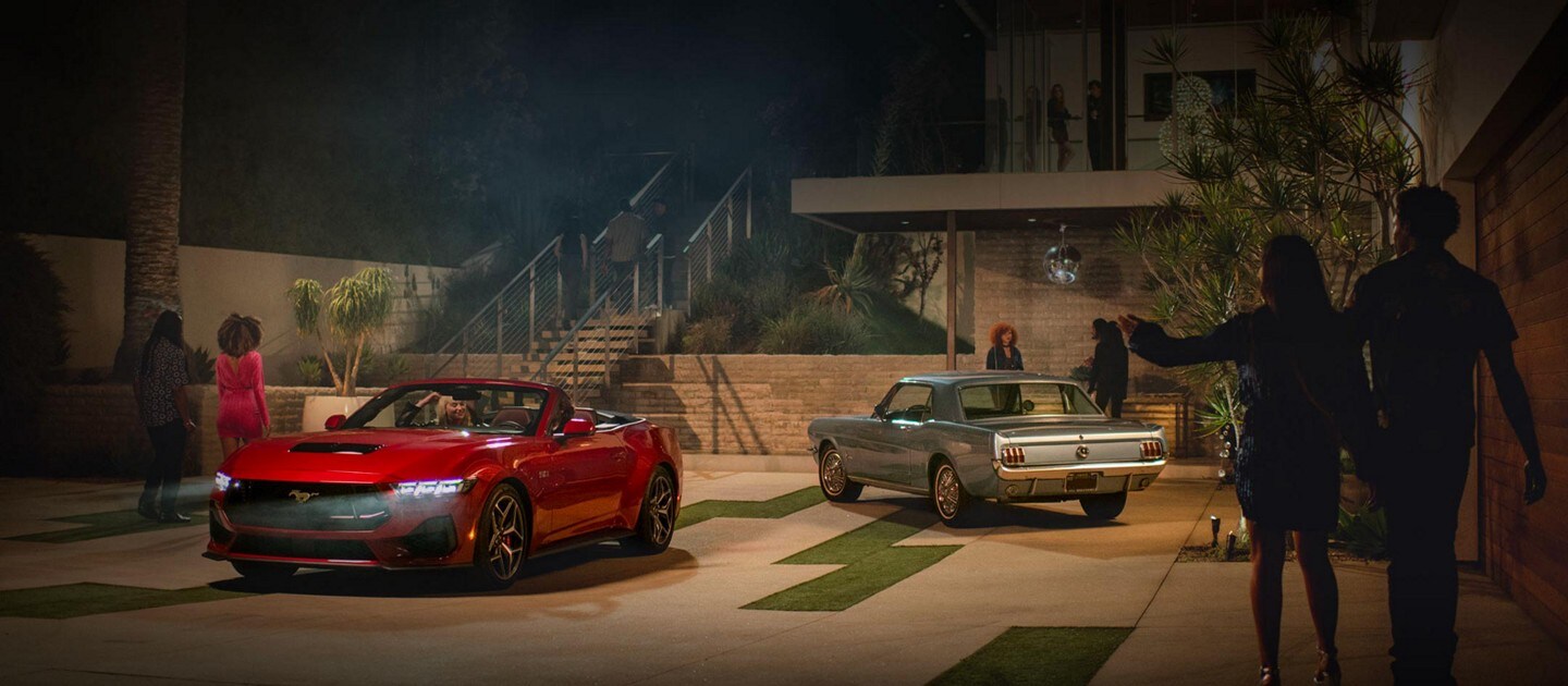 Un modelo Ford Mustang® GT 2024 convertible y un modelo Mustang de primera generación estacionados en una entrada con gente alrededor