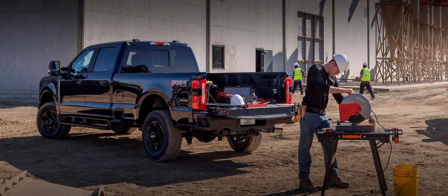 Hombre usando una sierra ingletadora en un lugar de trabajo con una camioneta Ford Super Duty® 2024 estacionada cerca con la puerta trasera baja