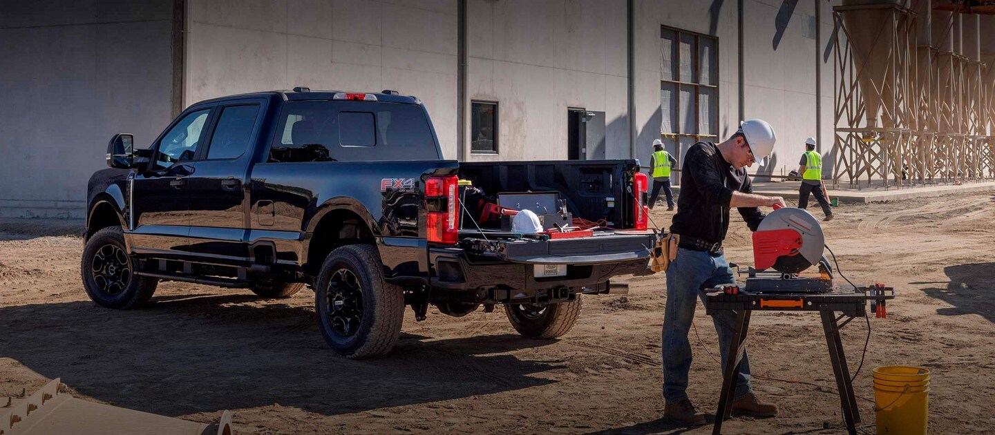 Man using a miter saw at a work site with a 2024 Ford Super Duty® truck parked nearby with tailgate down